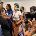 Foto de plateia de audiência pública promovida pelo Ministério da Educação mostra pessoas em pé, quietas. Uma delas, com a camiseta da entidade estudantil UBES, está com uma faixa sobre a boca, como uma mordaça. Divulgação/Humberto Costa
