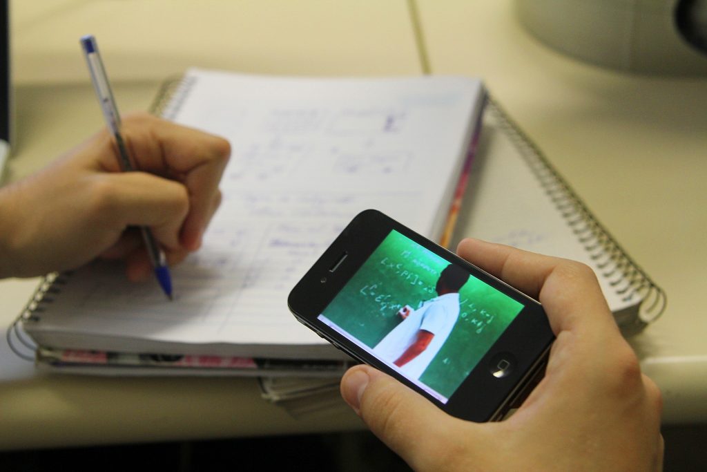 Estudante faz anotações em caderno enquanto assiste aula de matemática pelo celular