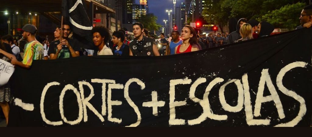 Jovens em um protesto com uma grande faiza preta escrito em branco: - cortes + escolas